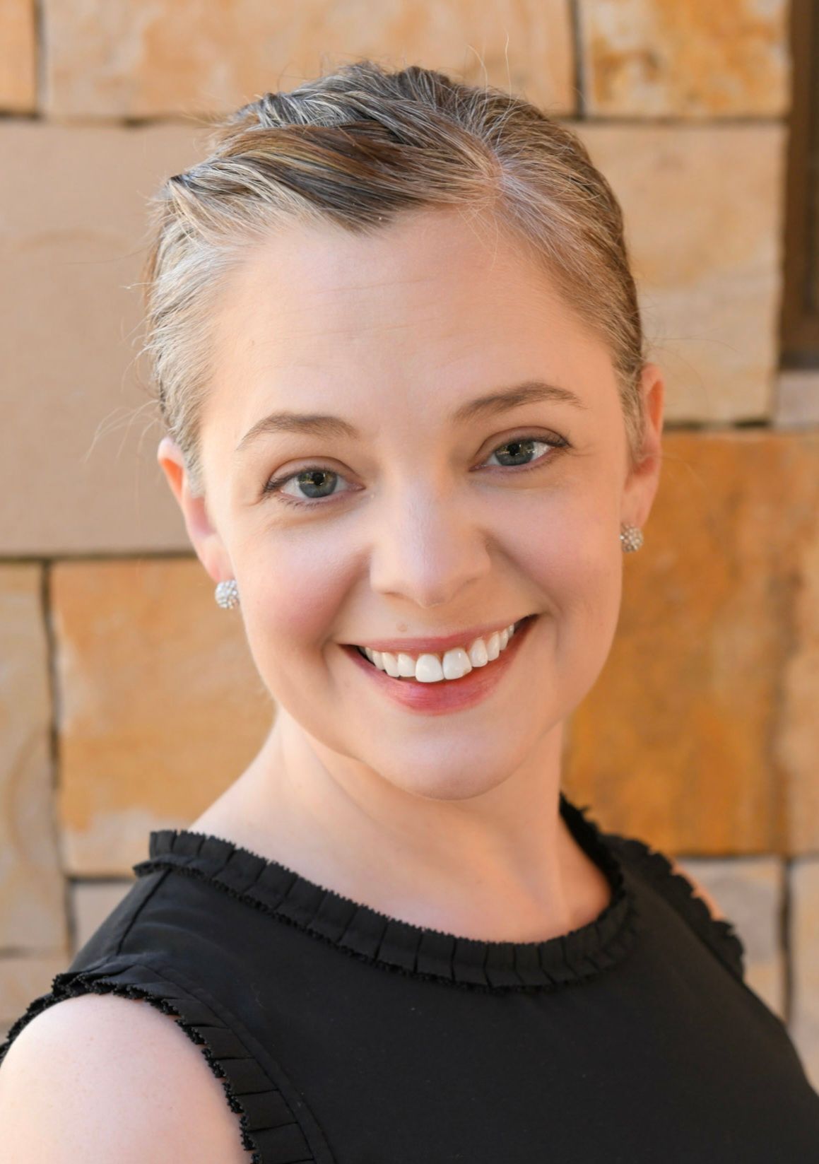 Woman smiling, wearing black top, against a stone wall background.