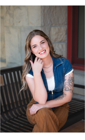 Woman with blonde hair smiles, standing in front of a floral backdrop; tattoo on arm.