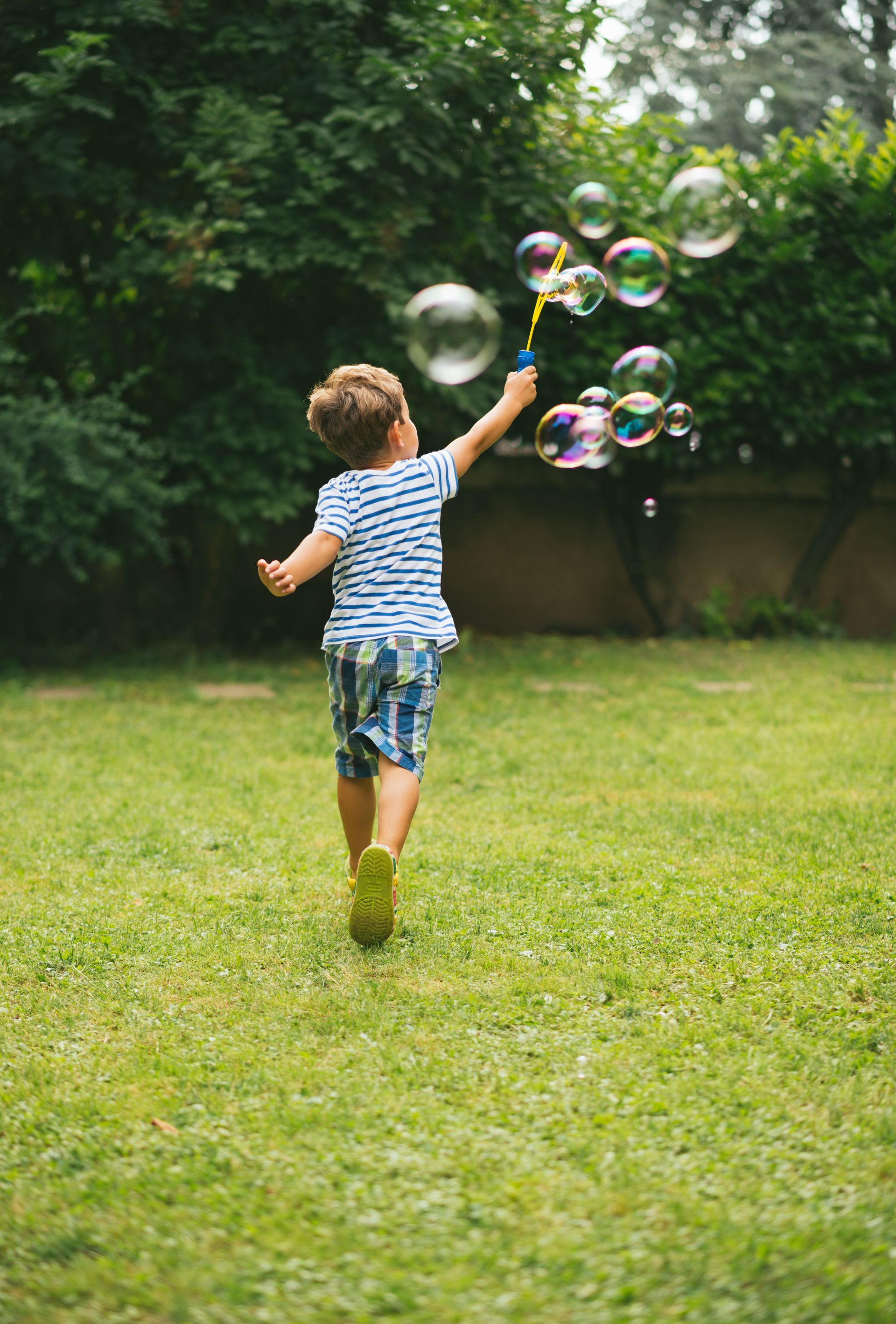 Boy in striped shirt and checkered shorts runs on grass, blowing bubbles in a backyard.
