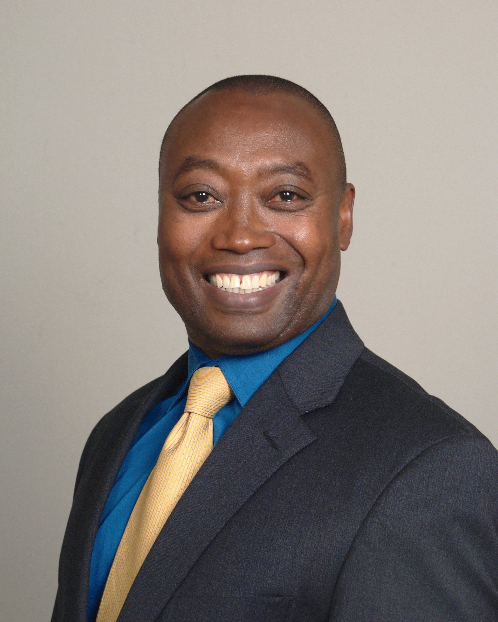 Smiling man in a suit, with a blue shirt and yellow tie against a gray background.
