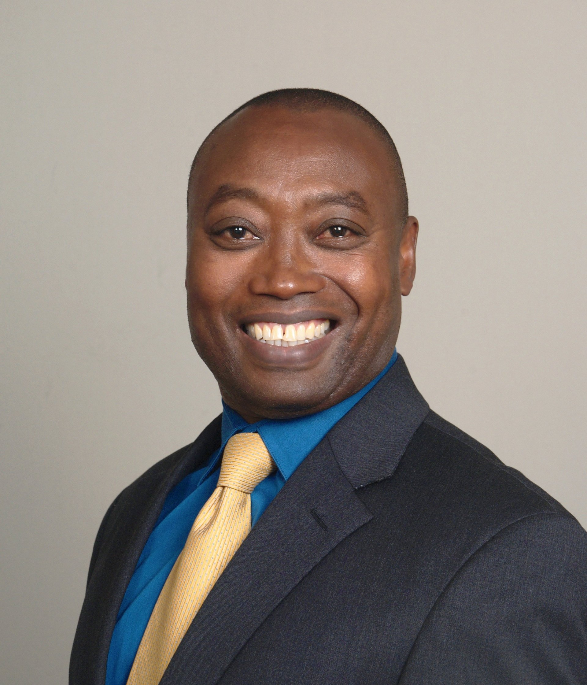 Man in blue shirt, yellow tie, and dark suit smiles against a gray background.