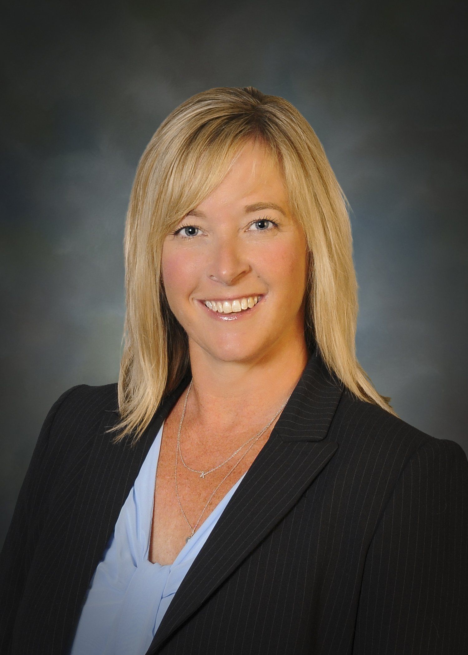 Woman in a dark blazer and light blue shirt smiles, with blonde hair and a necklace.