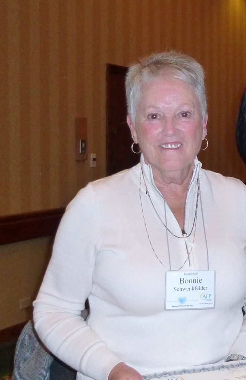 Woman with short hair, wearing a white shirt and name tag, smiles in a room.
