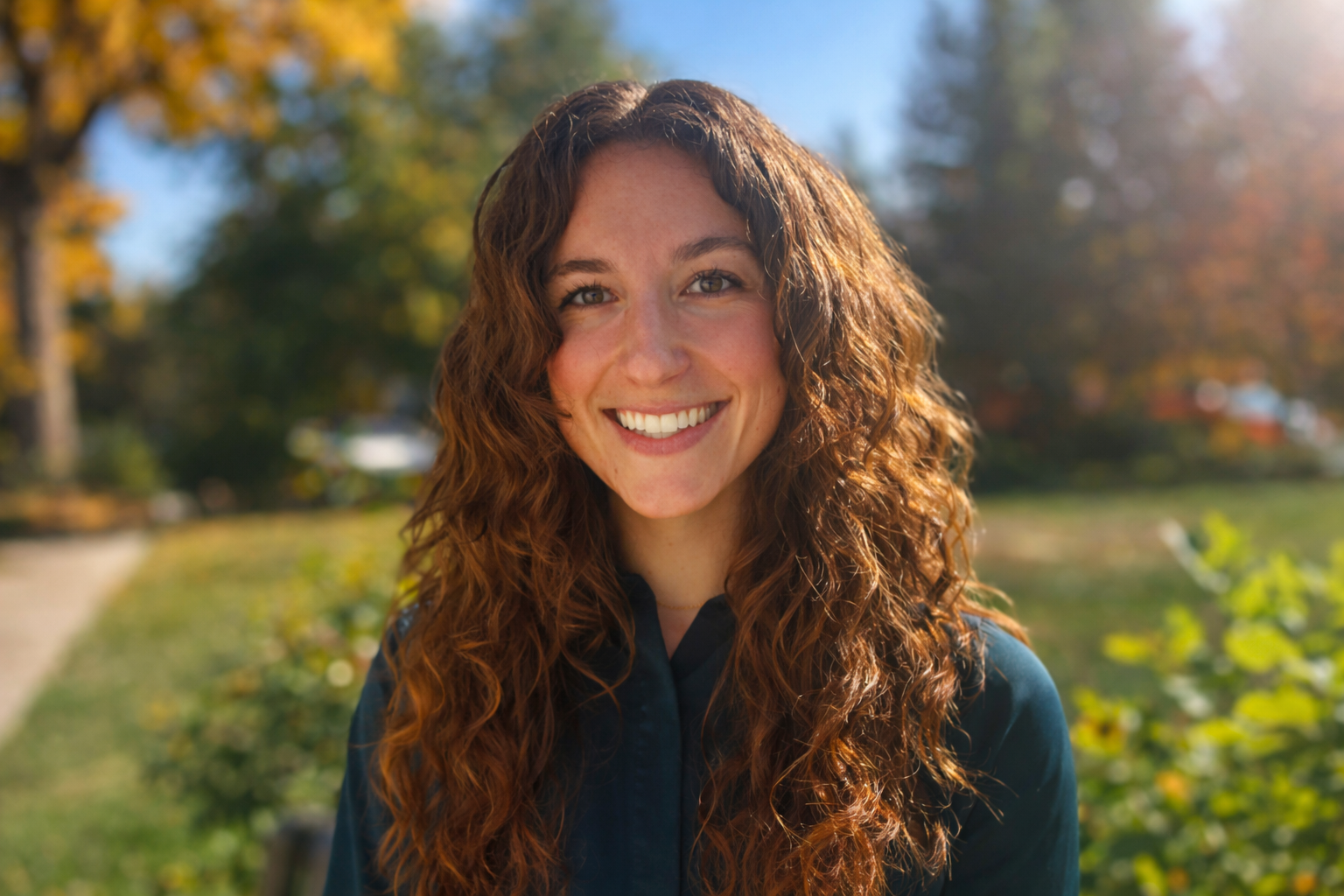 Woman with long, wavy brown hair smiles outdoors in front of green foliage and a tree.