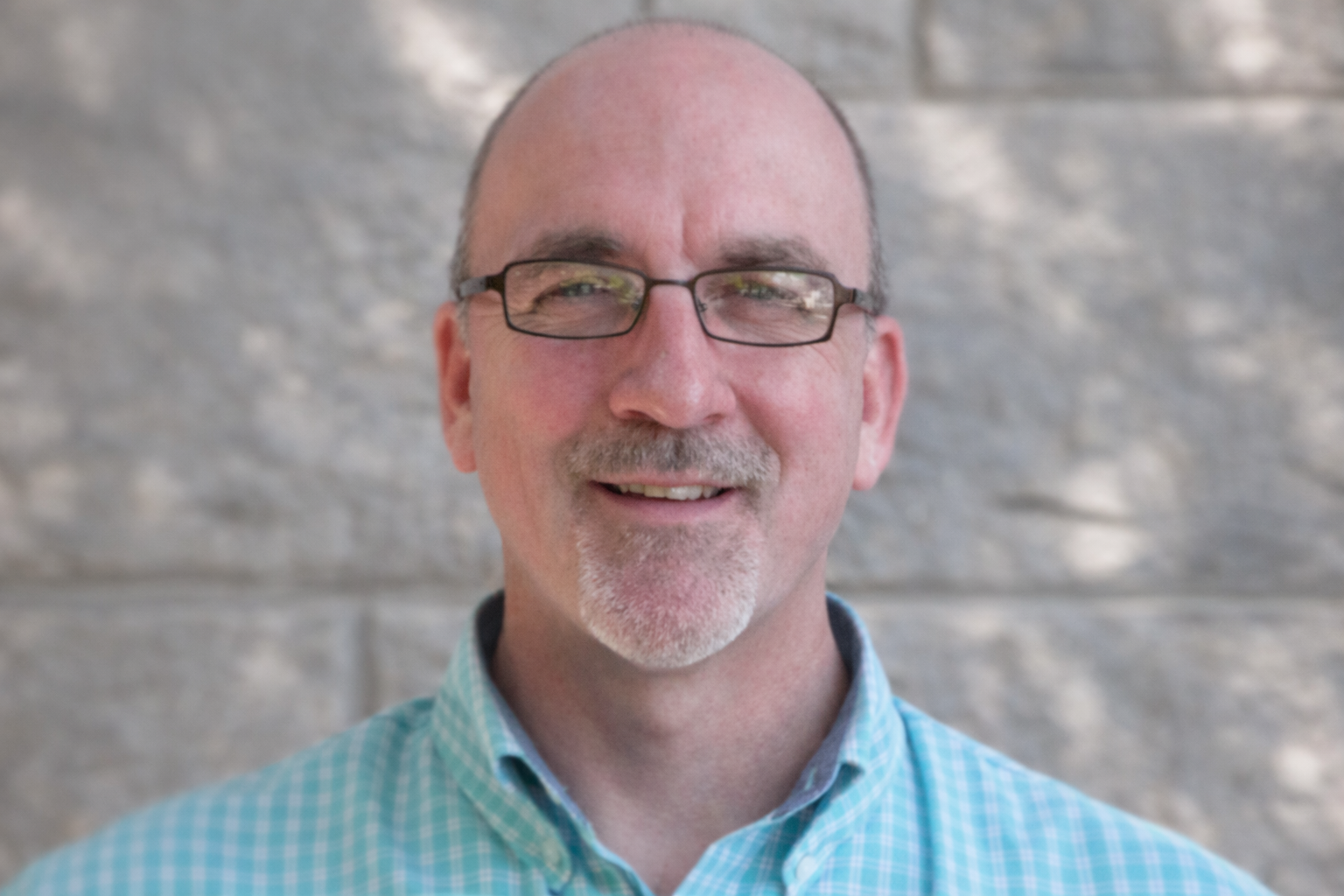 Man with glasses smiles, light blue shirt, in front of a gray brick wall.