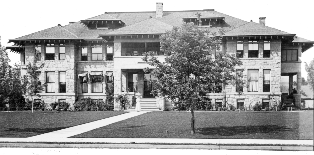 Large stone building with multiple windows, a porch, and a tiled roof, set back from a green lawn.