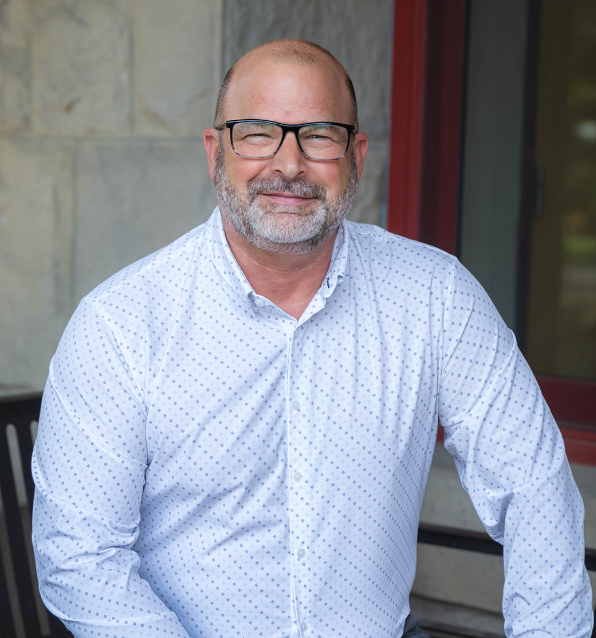 Man with a bald head and beard wearing a light pink shirt and burgundy tie, smiling.