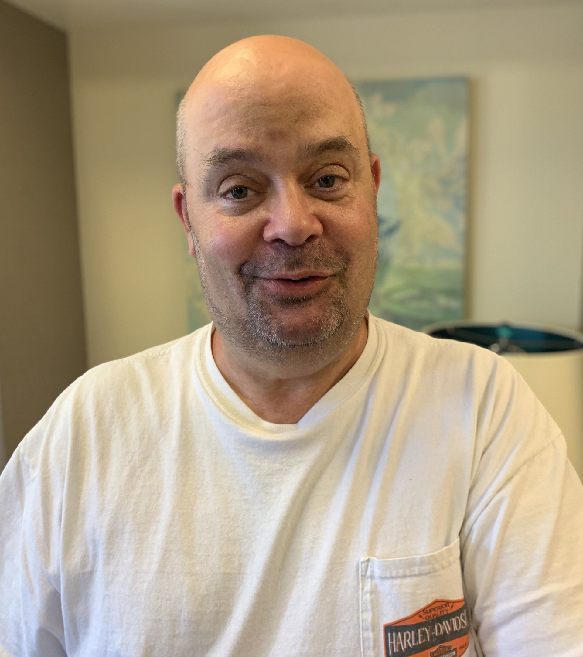 Bald man in a white t-shirt smiles, indoors. A Harley-Davidson logo on his shirt. Soft background.