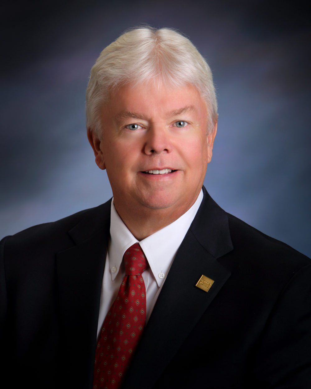 Man in a suit and red tie, smiling at the camera, against a blurred blue background.