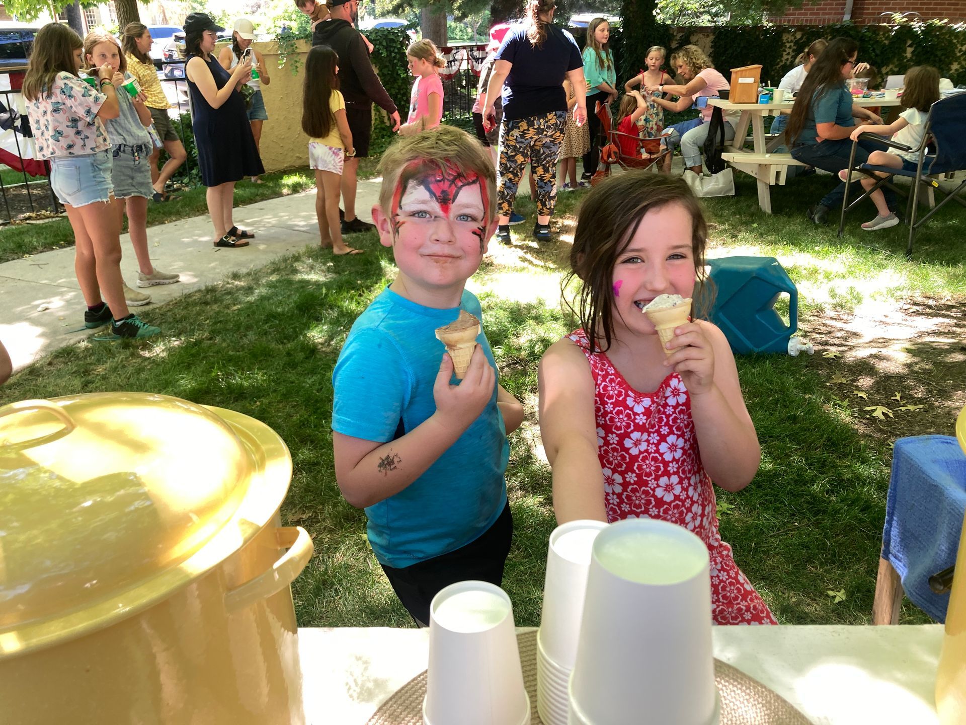Two children with face paint, eating ice cream cones at an outdoor event. People in background.