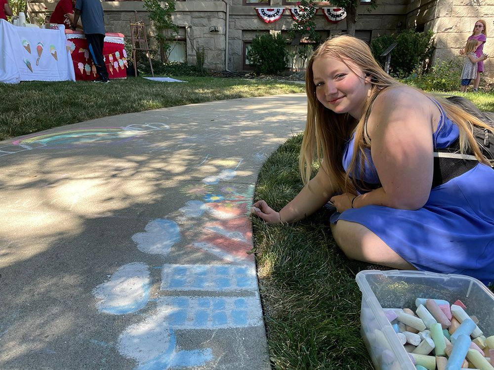 Woman drawing with chalk on a sidewalk, smiling. Chalk and artwork in progress. Outdoor event with decorations.