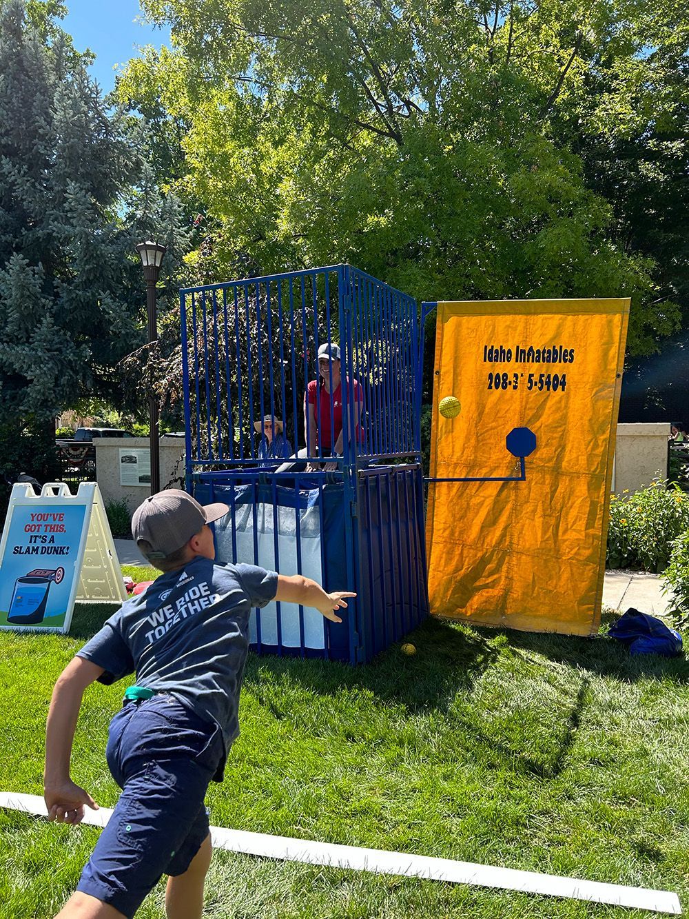 Boy throwing a ball at a dunk tank, aiming to dunk a person in the cage. Outdoors, sunny day.