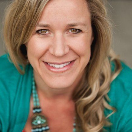 Woman with blonde hair, smiling, wearing a teal top and a beaded necklace.