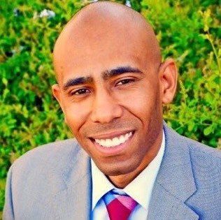 Man with bald head, smiling, wearing a gray suit, white shirt, and a pink tie, in front of greenery.