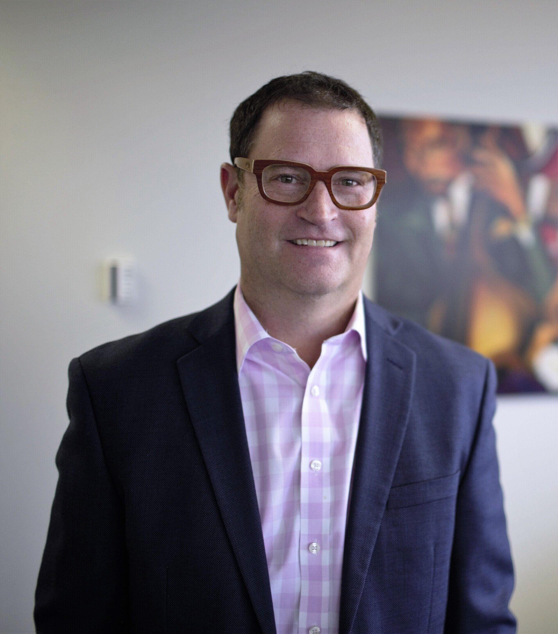 Man in a blue blazer and pink shirt, wearing glasses, smiling in an office setting.
