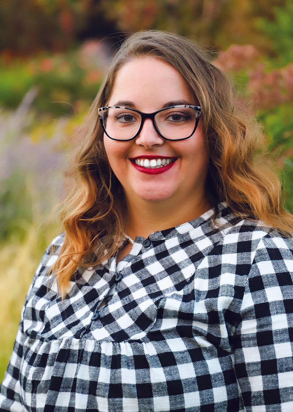 Woman with glasses, smiling in a black and white checkered shirt, outdoors.