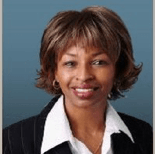 Woman with brown hair smiling, wearing a black blazer and white shirt. Blue background.