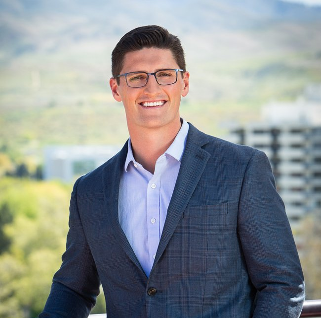 Man in blue blazer and glasses smiles outdoors.