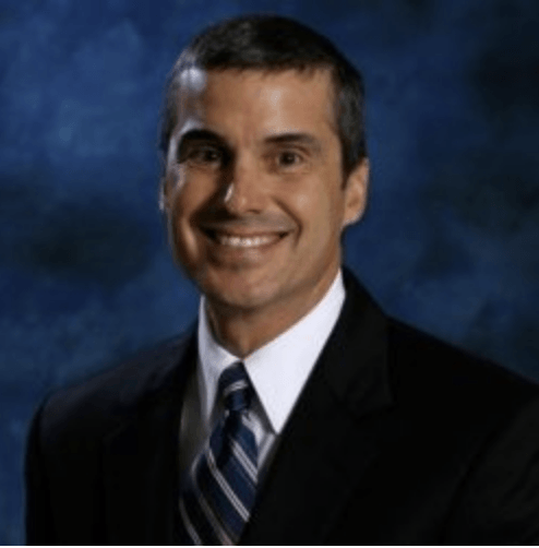 Man in a suit smiles at the camera against a blurred blue background.