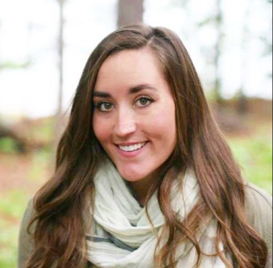 Woman with long brown hair smiling, wearing a light scarf outdoors in front of blurred trees.