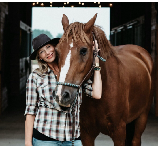 Woman in plaid shirt and hat with brown horse in barn.
