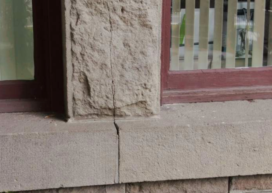 Close-up of a cracked stone windowsill and adjacent column, near a window with a burgundy frame.