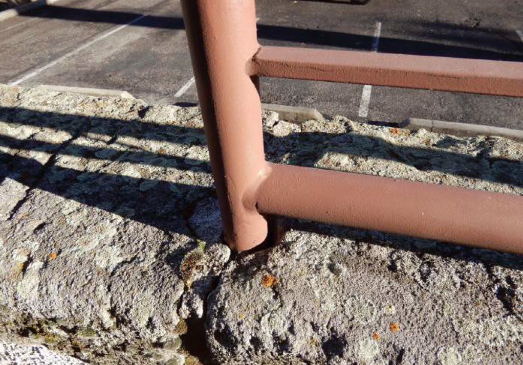 Brown metal railing attached to a textured, light gray stone wall, with parking lot in the background.