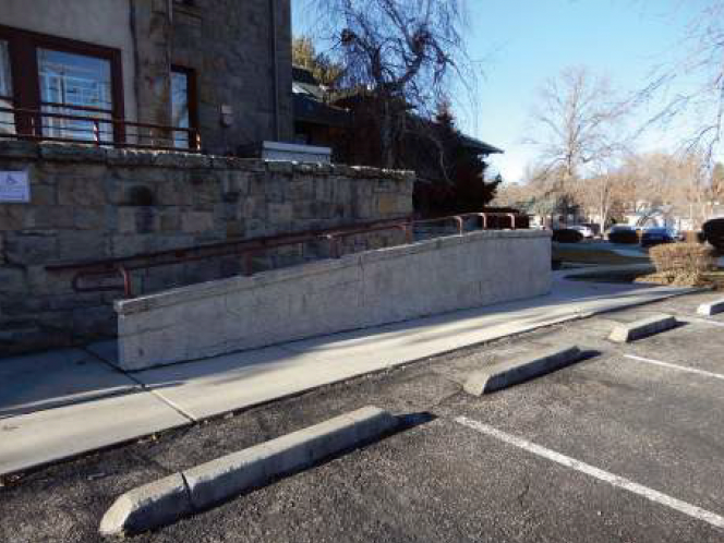 Concrete wheelchair ramp next to a stone building in a parking lot.