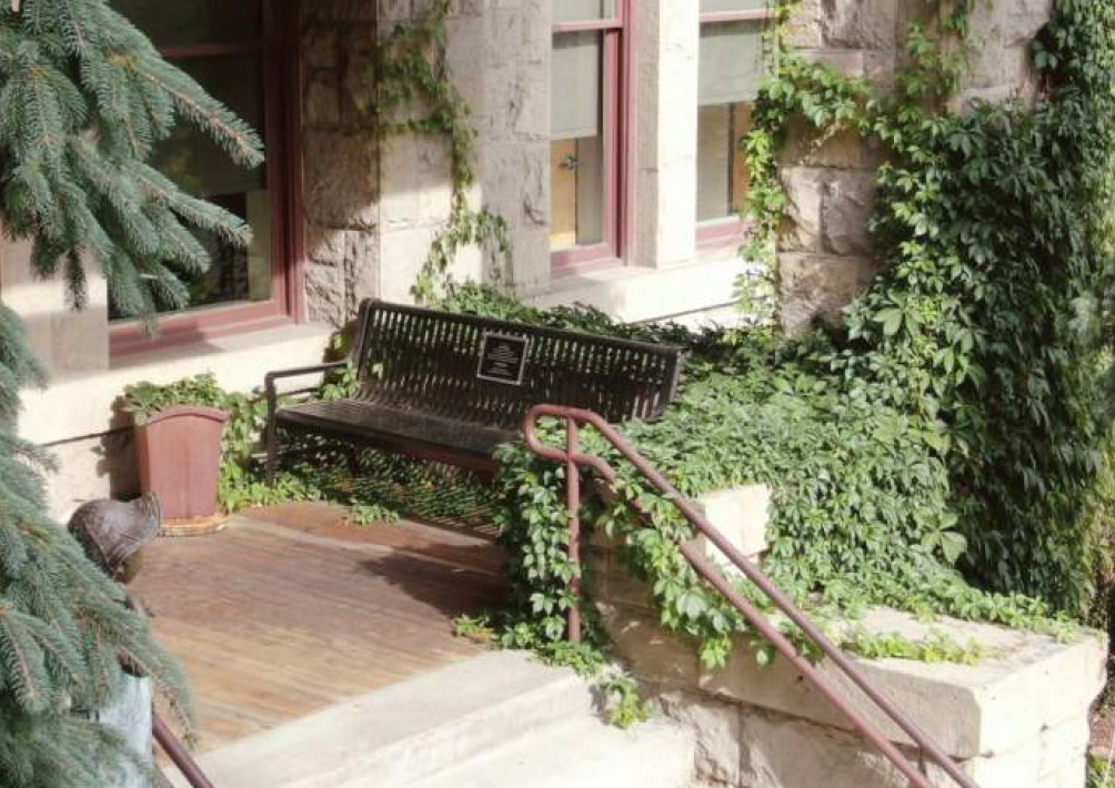 Wooden bench on a porch, surrounded by green foliage, next to a building with stone walls and windows.