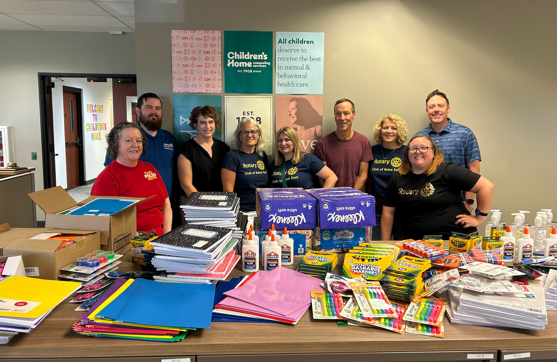 Group of people with school supplies on a table; gathered for donation.