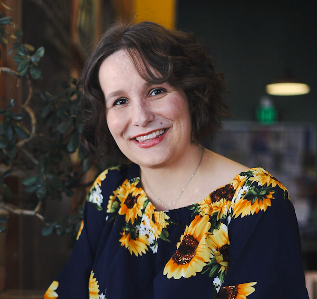 Woman smiling, wearing a navy blouse with sunflower print, inside a building.