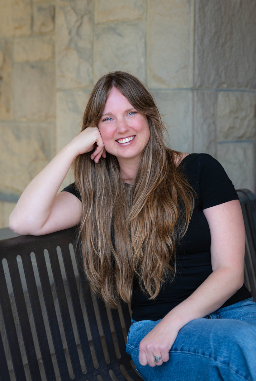Woman with long blonde hair smiles, wearing a black and white patterned shirt, seated in a room with blue wall.