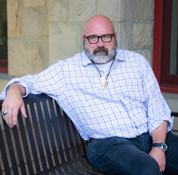 Bald man with glasses and beard, wearing a light blue checked shirt and a pendant, smiling.