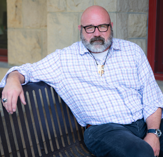 Man with glasses and beard wearing a checked shirt and bone pendant. Sitting indoors with patterned background.