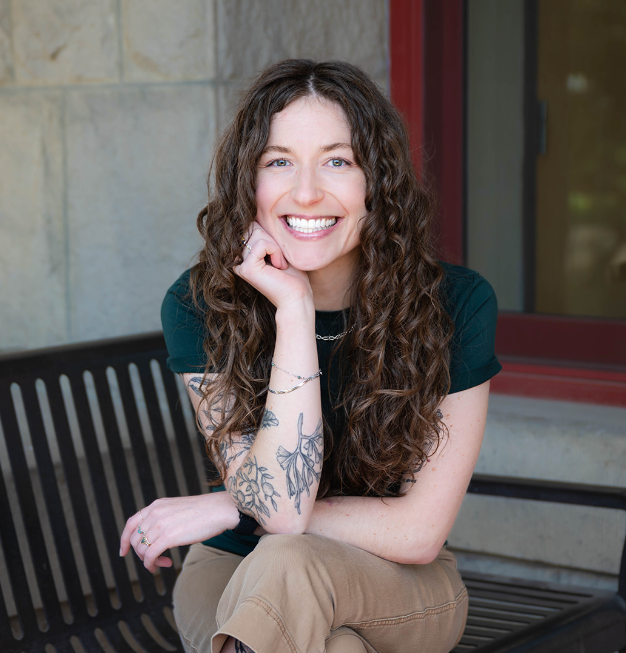 Woman with long, wavy brown hair smiles outdoors in front of green foliage and a tree.