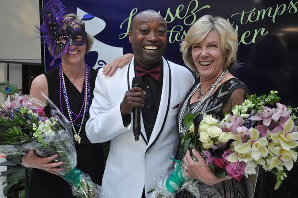 Three people smiling, holding bouquets, at an event. Man in white suit, women with masks and necklaces.