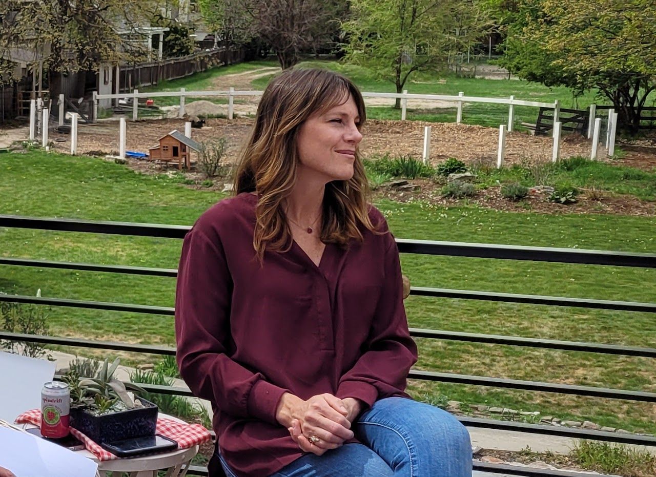 Woman in burgundy shirt sits outside, smiling, with a fenced yard and trees in the background.