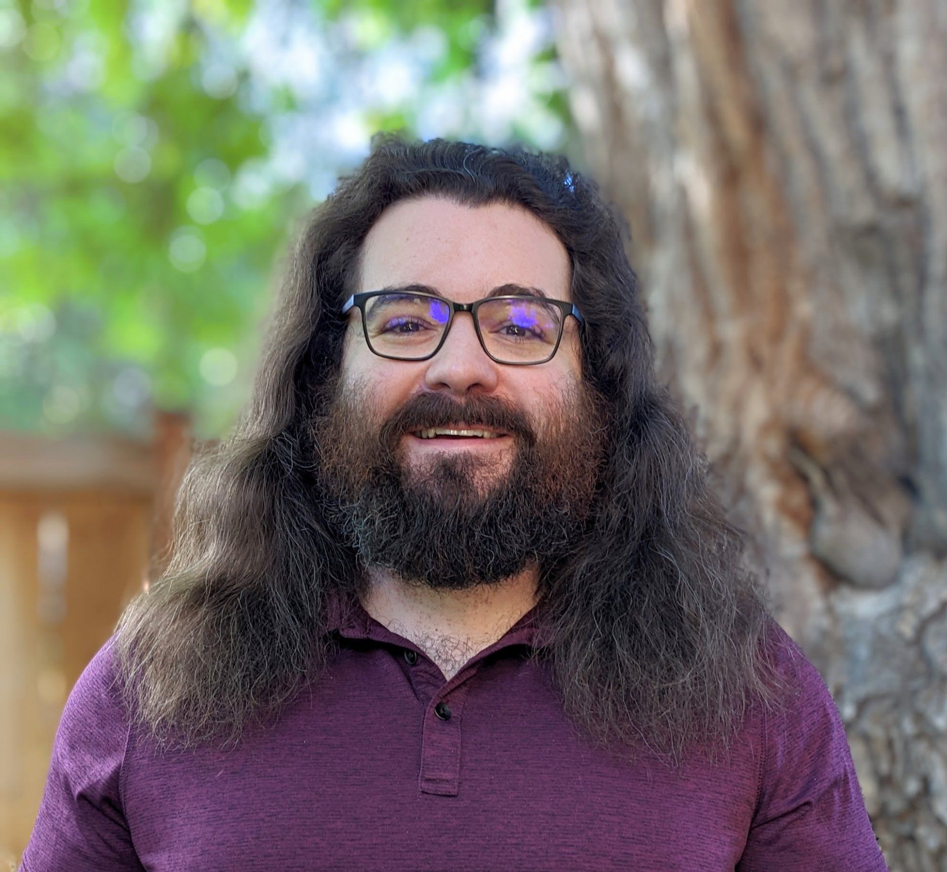 Man with glasses and long brown hair smiles, wearing a purple shirt, set outdoors with a tree.