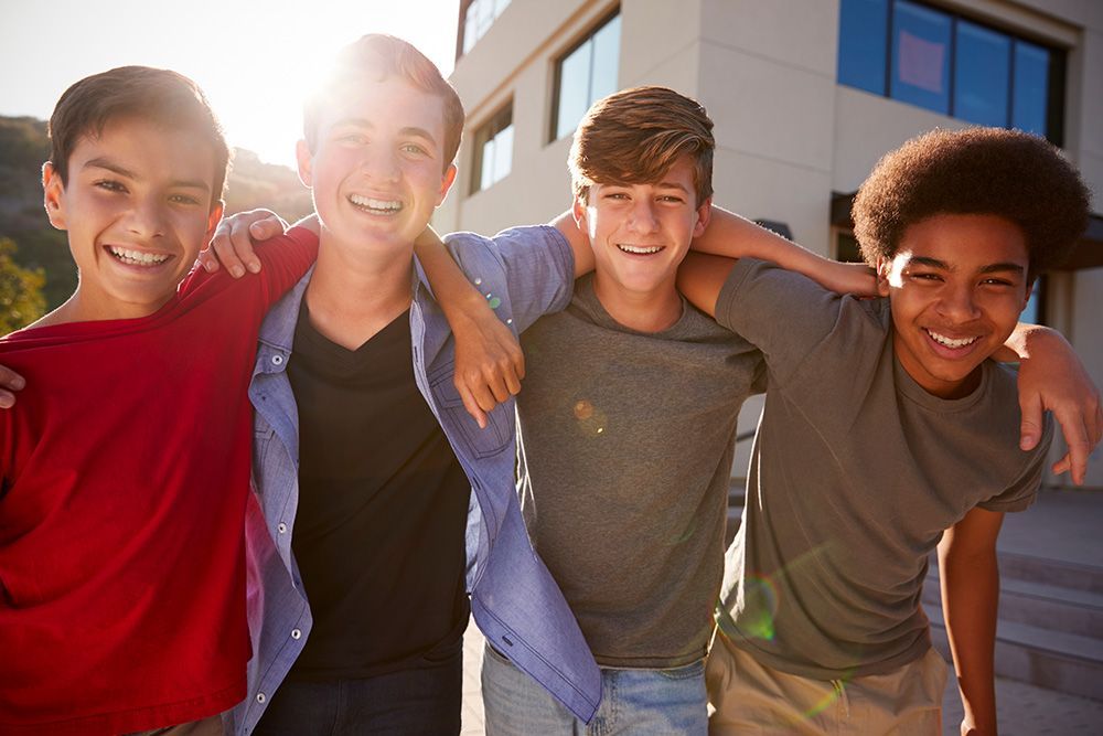 Four smiling teens with arms around each other pose outside a building on a sunny day.