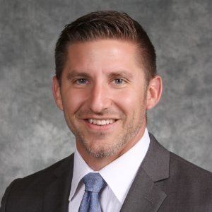 Man in suit smiles at the camera, against a mottled gray background.