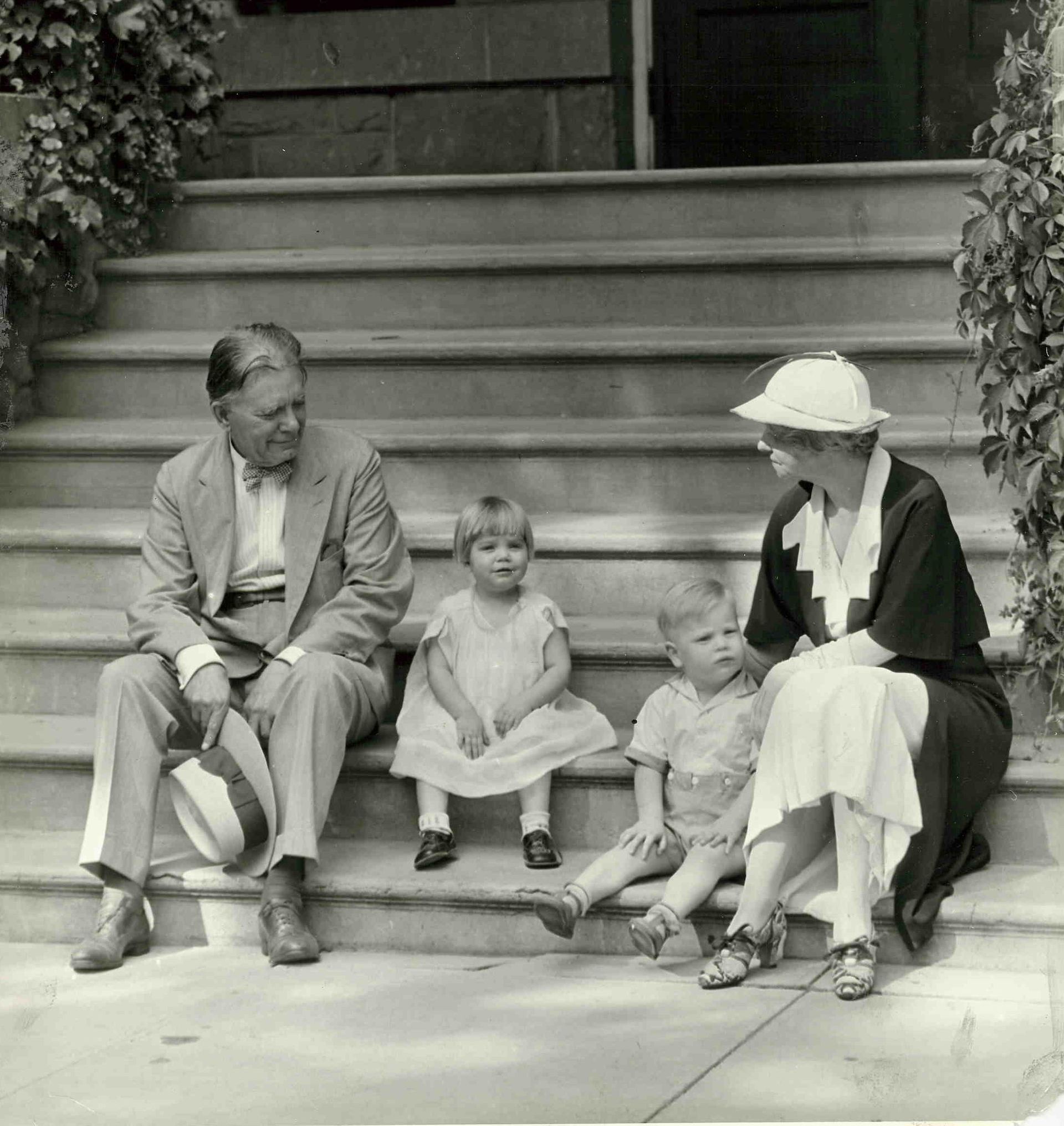 A family sits on outdoor steps. A man holds a hat, a woman wears a hat, and two children sit between them.