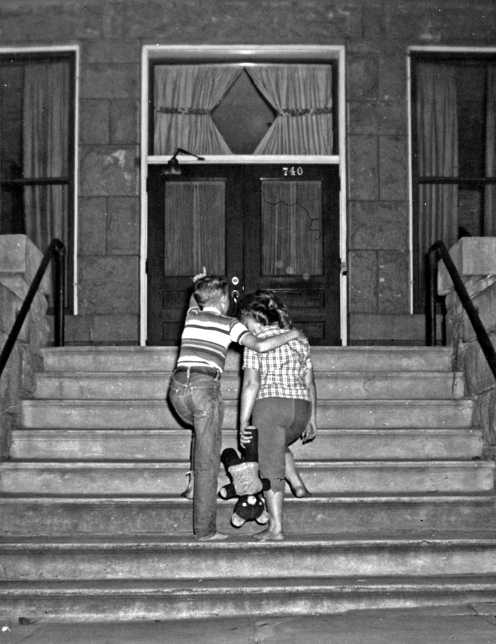 Two children climbing stone steps towards a building's front door; a teddy bear sits on a step.