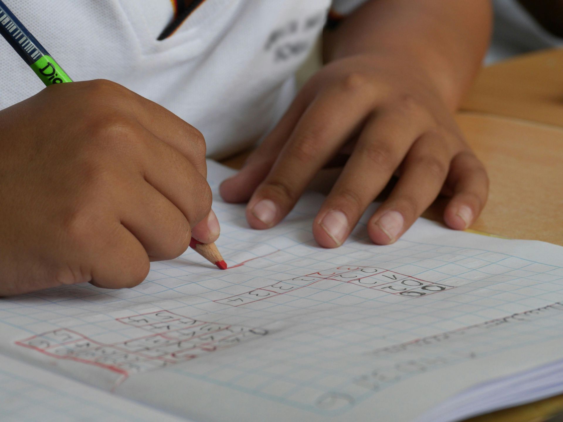 Child's hands writing with a red pencil on graph paper, close-up.