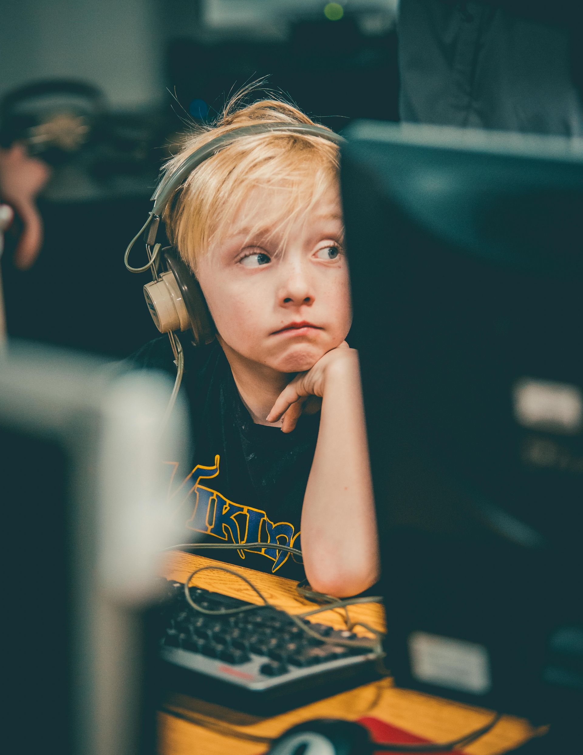Boy with blonde hair, wearing headphones, looks perplexed while sitting at a computer.
