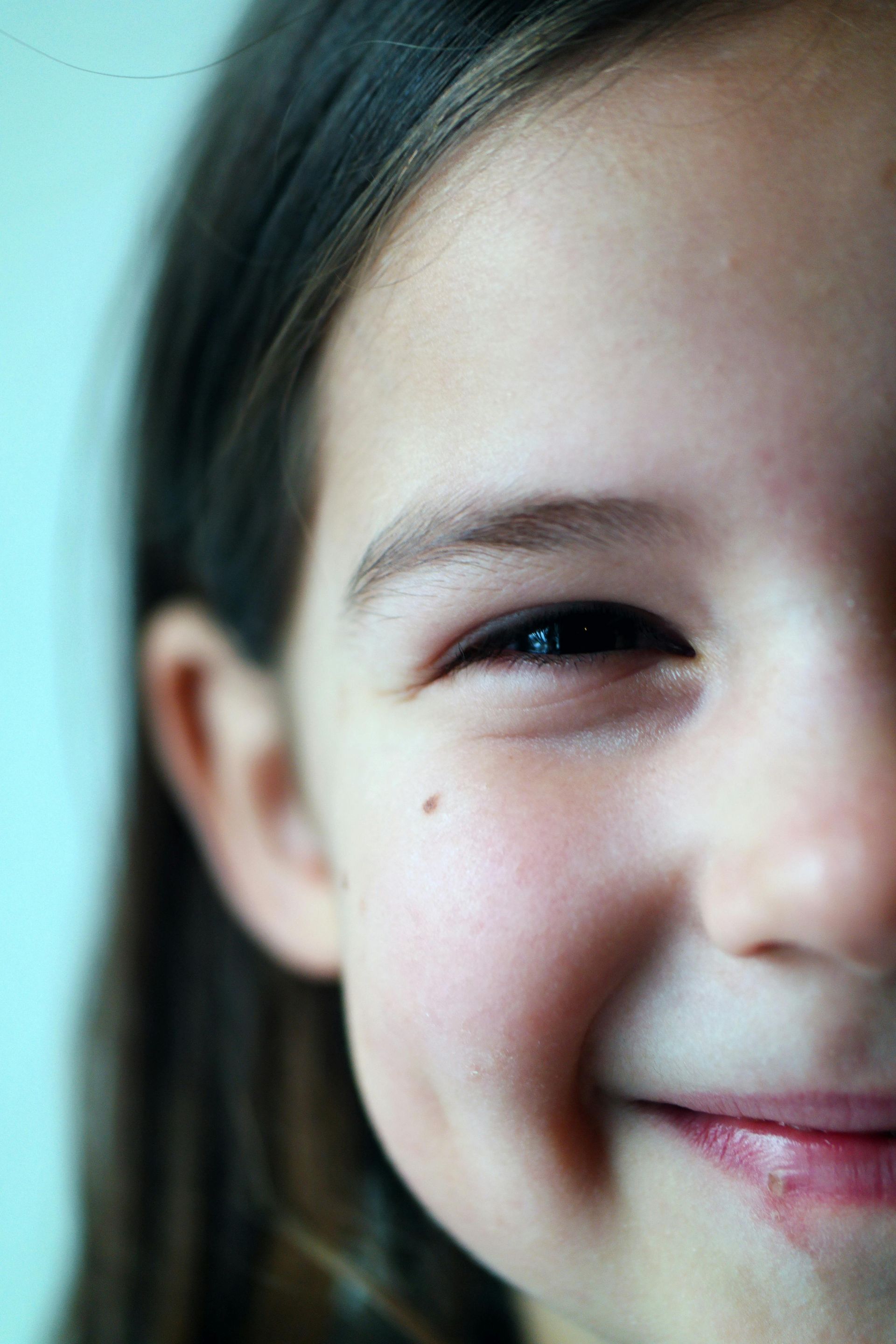 Close-up of a smiling child's face; one eye visible, dimple on cheek, light skin, brown hair.