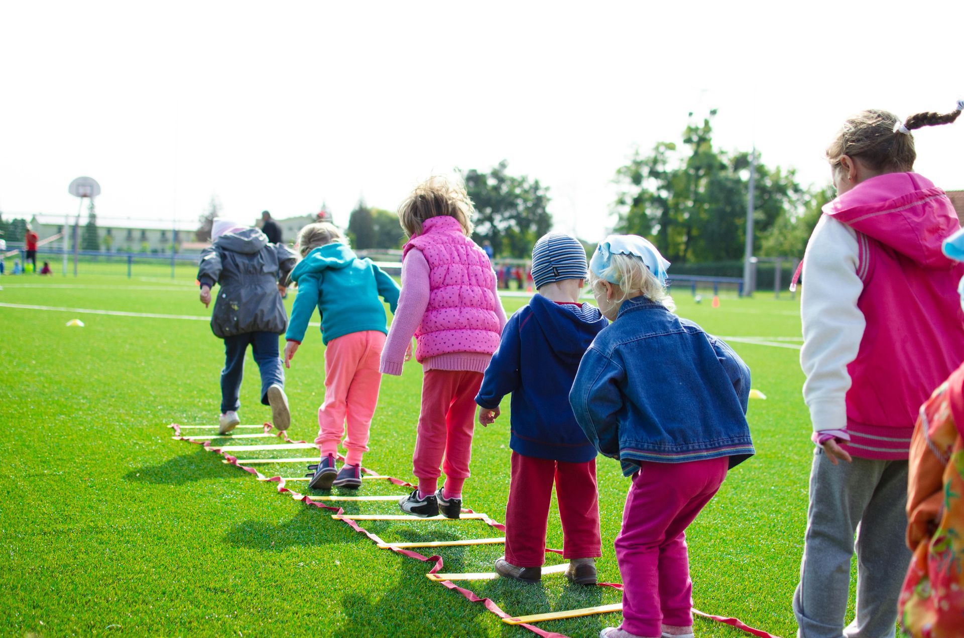 Children playing on an agility ladder on a green sports field.