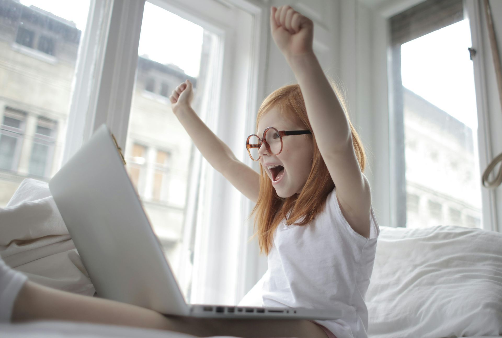 Girl with red hair and glasses cheers with arms raised while using a laptop on a bed by a window.