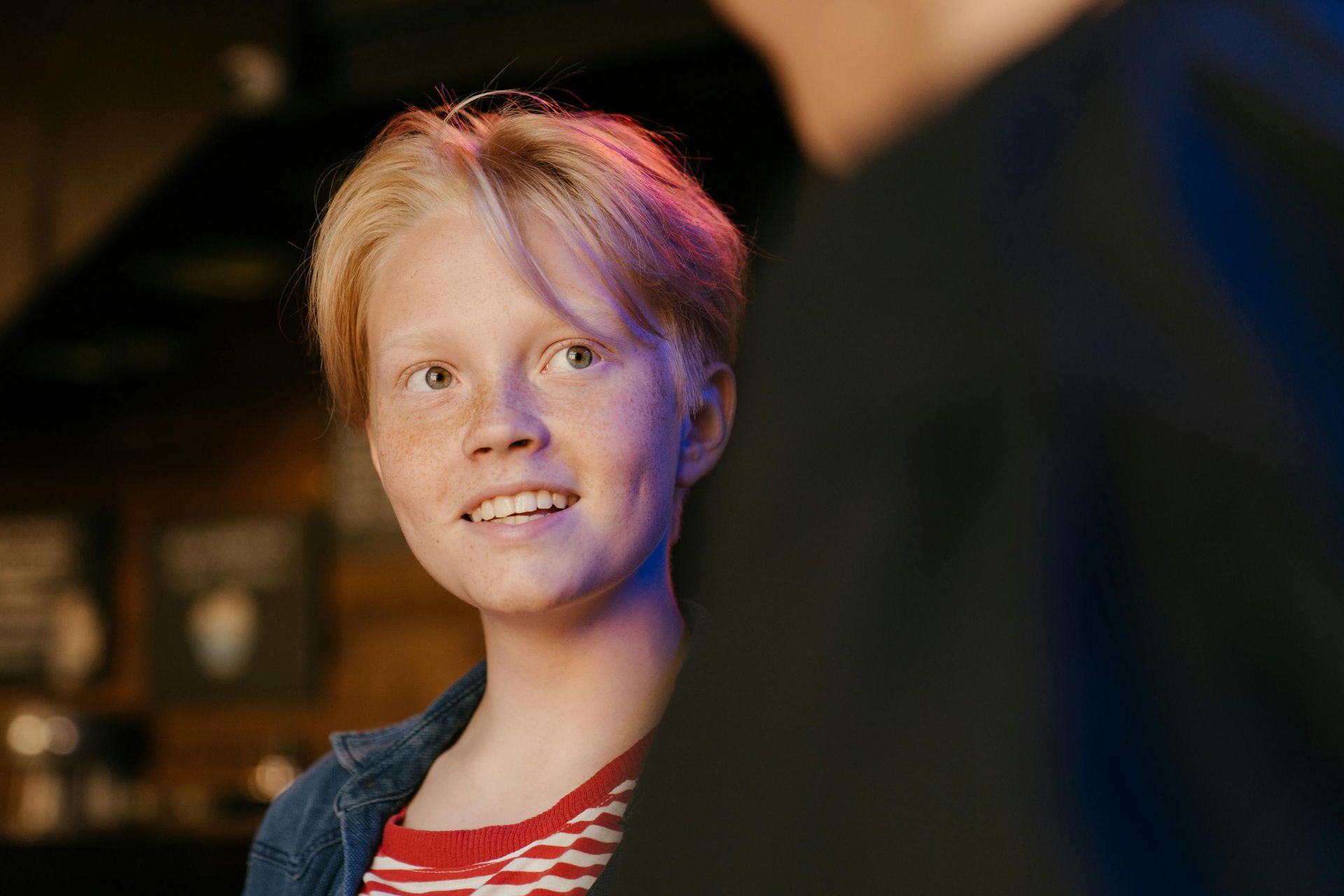 Blonde person with freckles smiles and looks to the side, next to a person in black clothing.