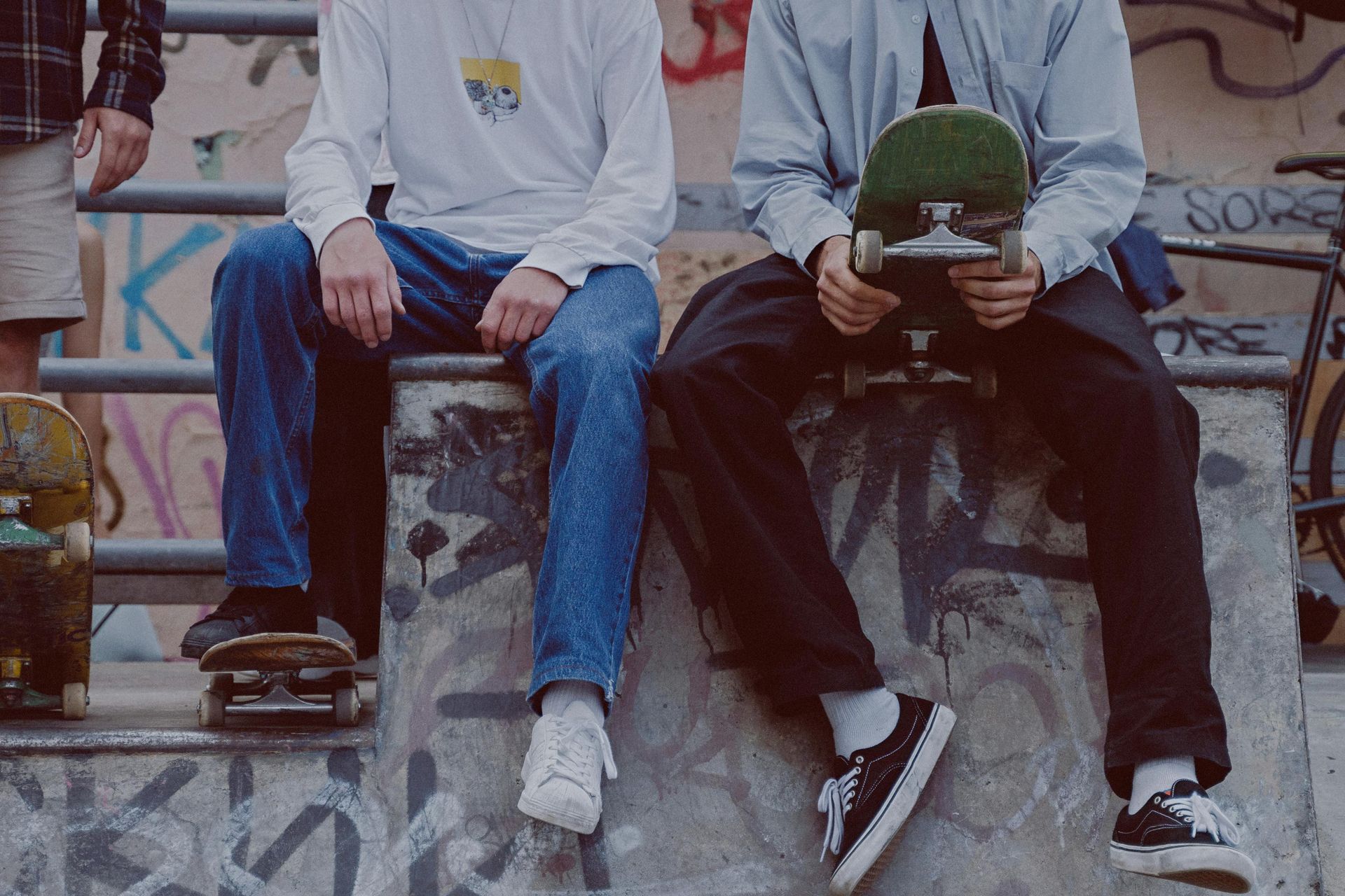Three people sitting on a skate park ledge holding skateboards. Concrete and graffiti background.