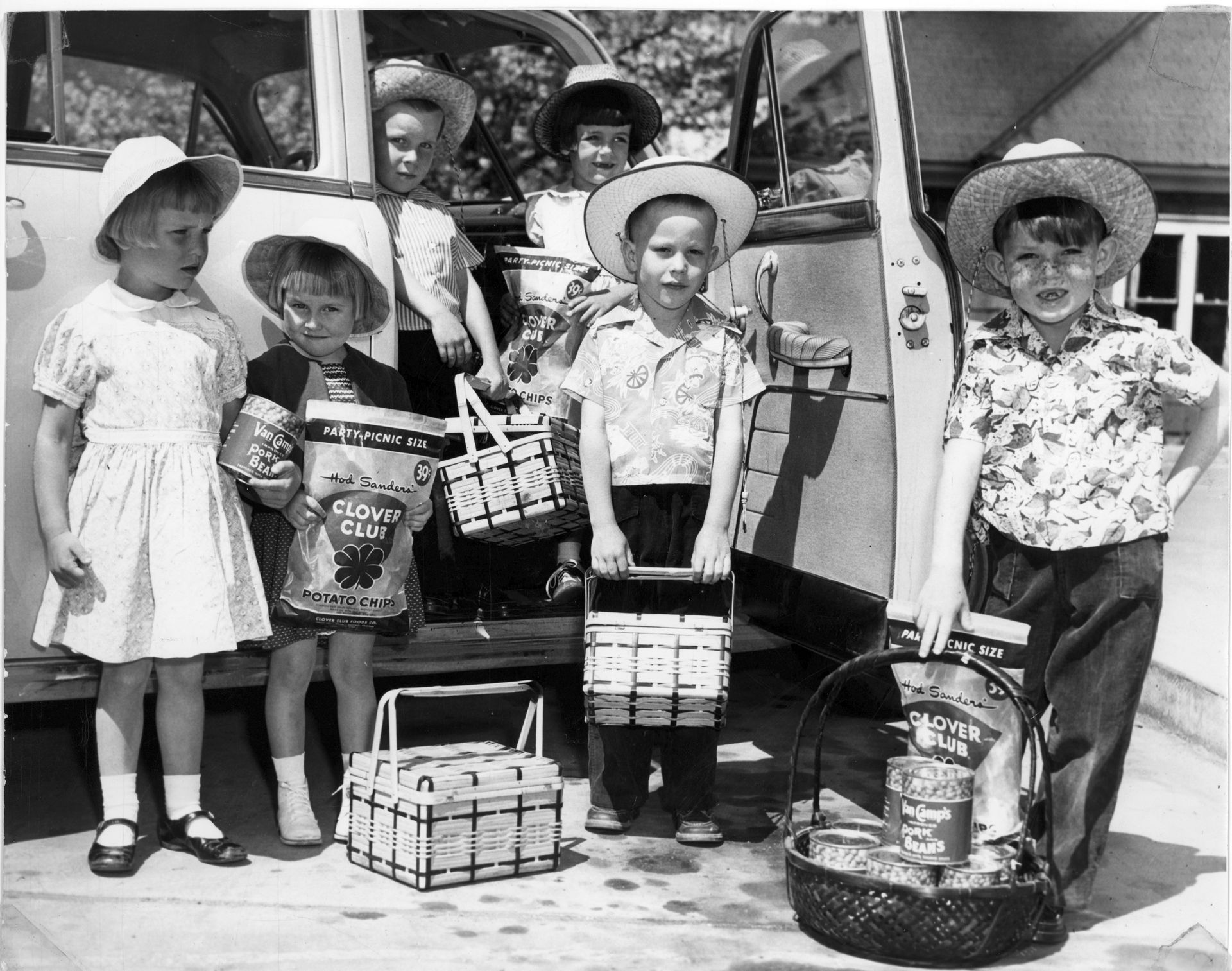 Children in hats and summer clothes, holding picnic baskets and bags by an old car.